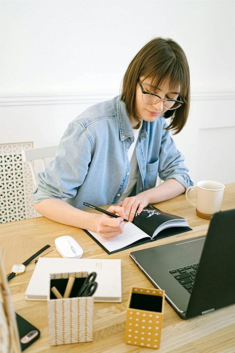 Woman in glasses writing in notebook at home office desk with laptop and coffee.